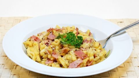 Käsespätzle mit Schinken und Petersilie in einer weißen Schüssel, auf einem natürlichen Tischuntergrund serviert.