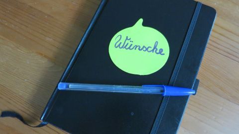Um Stress mit Geschenken zu vermeiden, kannst du ein Buch mit allen beiläufig erwähnten Wünschen deiner Lieben führen! Ein schwarzes Notizbuch mit einem grünen Haftnotizzettel und einem blauen Stift liegt auf einem Holztisch.