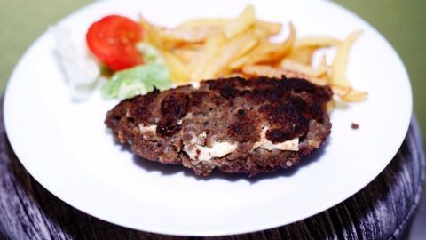 Ein gegrilltes Bifteki liegt auf einem weißen Teller, begleitet von Pommes und einem frischen Salat mit Tomate.