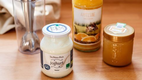 Ein Glas Joghurt steht auf einem Holztisch, während eine Flasche Saft und ein Honigglas daneben liegen.