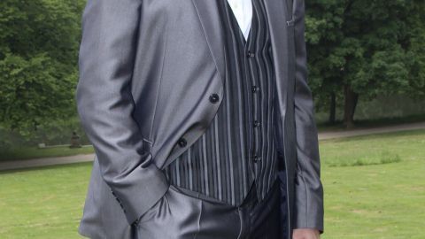 Ein eleganter Mann in einem grauen Anzug mit Weste steht lässig im Freien, umgeben von grüner Wiese und Bäumen.