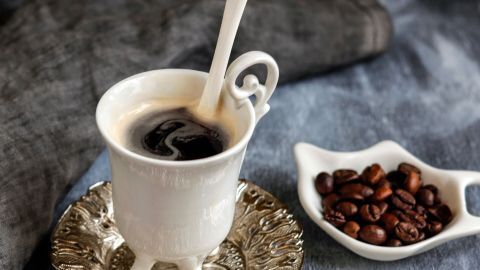 Kaffee nach Art des wilden Westens: Ein Tipp zum Schmunzeln, zum Kochen von starkem Kaffee. Eine elegante Tasse Kaffee steht auf einem filigranen Untersetzer, während eine kleine Schale mit Kaffeebohnen daneben liegt.