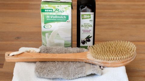 Erlebe Luxus wie Cleopatra mit einem Milchbad und Olivenöl für samtweiche Haut. Mit einer Bürste kannst du deine Haut sanft massieren. Ein einfaches Wellness-Erlebnis für Zuhause. Eine Holzbürste liegt auf Frotteehandtüchern neben H-Vollmilch und nativem Olivenöl auf einem Holztisch.