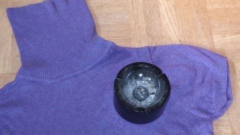 Ein lila Pullover liegt auf einem Holzfußboden, während ein schwarzes Gerät zur Rauchgeruchsbeseitigung darauf platziert ist.
