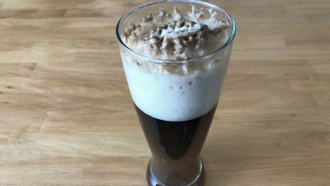 Ein Glas Goasmaß steht auf einem Holztisch, gefüllt mit dunklem Getränk und einer schaumigen, karamellisierten Haube.