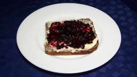 Eine Scheibe Brot ist mit schwarzer Johannisbeermarmelade belegt und auf einem weißen Teller vor einem blauen Tischcloth platziert.