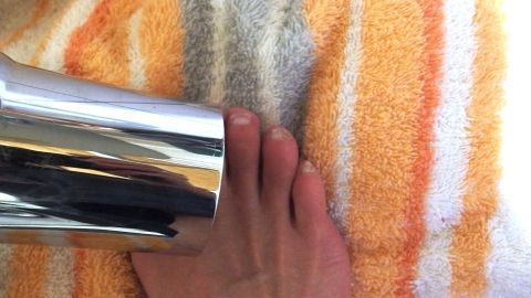 Dieser Tipp zum Vorbeugen von Fußpilz wirkt 100-%ig: Zehenzwischenräume nach der Dusche oder dem Bad föhnen. Ein Haartrockner föhnt einen Fuß auf einem gestreiften Handtuch, um Fußpilzprophylaxe zu unterstützen.