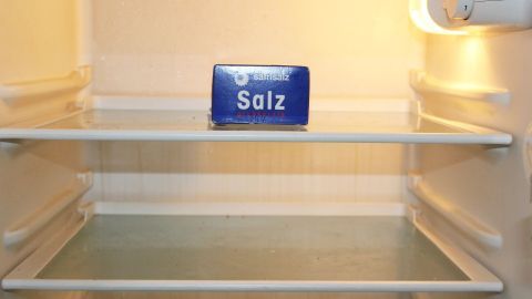 Mithilfe von Salz lässt sich Schimmelbildung in einem leeren Kühlschrank vermeiden. Ein leeres Kühlschrankregal zeigt eine Verpackung mit dem Wort "Salz", um Schimmel im Kühlschrank zu vermeiden.