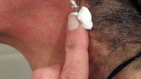 Flecken von Haarfarbe auf der Haut kann man mit Zahnpasta entfernen. Eine Hand mit Zahnpasta berührt die Haut hinter dem Ohr, um Flecken nach dem Haarefärben zu entfernen.