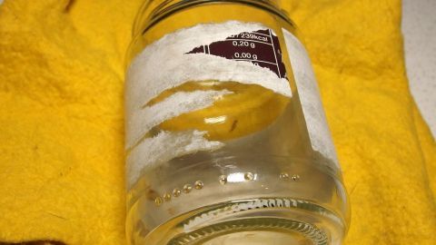 Ein Glas mit Kleberesten liegt auf einem gelben Tuch, während ein Orangenreiniger verwendet wird, um die Etiketten zu entfernen.