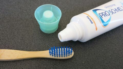 30 % weniger Karies: Das kann man erreichen, in dem man nach dem Zähneputzen den Mund nicht mit Wasser ausspült. Eine Zahnbürste liegt neben einer Zahnpasta, die Karies verhindern soll, auf einem Tisch mit grauer Oberfläche.