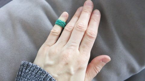 Eine Hand mit einem grünen Ring liegt auf einem grauen Stoff. Sie scheint entspannt und ruhig zu sein und vermittelt eine friedliche Atmosphäre.