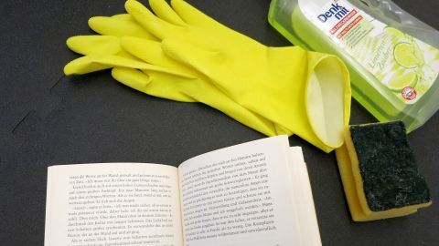 Gelbe Gummihandschuhe liegen neben einem Schwamm, Reinigerflasche und einem geöffneten Buch auf schwarzem Hintergrund.