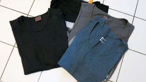T-Shirts liegen gefaltet auf weißen Fliesen, schwarz, grau, blau und mit Etiketten sichtbar.