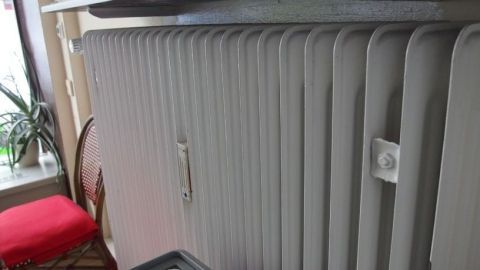 Ein Heizkörper ist am Fenster montiert und gibt Wärme ab, während ein roter Stuhl im Hintergrund steht.