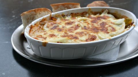 Gyros in Metaxasoße mit Käse überbacken wird heiß serviert in einer weißen Auflaufform, umgeben von knusprigem Brot.