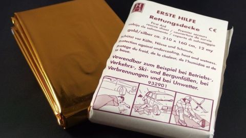 Die Rettungsdecke liegt zusammen mit einer Verpackung, die Anweisungen zur Nutzung in Notfällen gibt, auf schwarzem Untergrund.