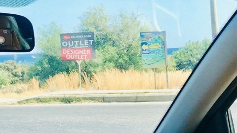 Extra zu einem Outlet mit dem Auto zu fahren ist ziemlich teuer. Wenn man in Urlaub fährt, kann man auch noch einen Abstecher machen. Das spart Geld und Zeit. Ein Auto fährt vorbei an einem Schild mit der Aufschrift „OUTLET DISCOUNT“, umgeben von trockener Landschaft.