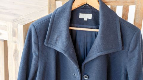 Hochwertige Kleidung wie dieser elegante Mantel hält länger und spart auf Dauer Geld. Investiere in Qualität, um weniger häufig ersetzen zu müssen — so profitierst du von besserem Tragekomfort und einem zeitlosen Stil. Ein hochwertiger Mantel hängt an einem Holzstuhl in einem hellen Raum. Keine Textinformationen sichtbar.
