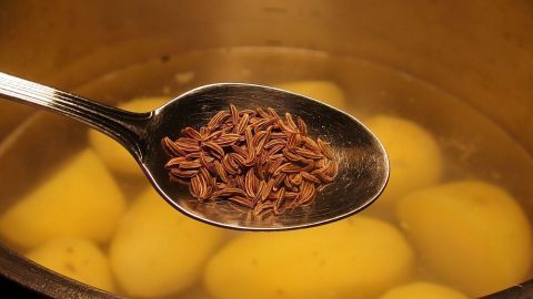 Salzkartoffeln mit etwas Kümmel kochen: schmeckt lecker und Kartoffeln bleiben länger frisch. Auf einem Löffel liegt Kümmel, während daneben Salzkartoffeln in einem Topf kochen. Die Küche ist warm und einladend.