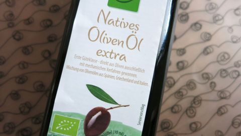 Eine Flasche mit nativem Olivenöl steht auf einem Tisch, umgeben von einem dezenten, gemusterten Hintergrund.