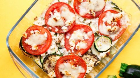 Ein Zucchini-Tomaten-Gratin mit Feta in einer Glasform, überbacken und auf einem gelben Tisch serviert.