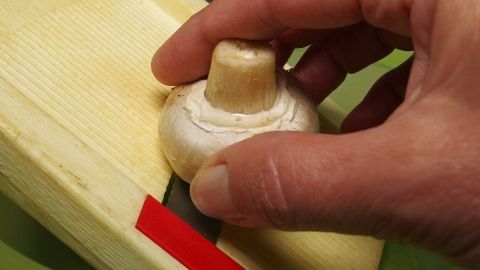 Feste Champignons blitzschnell mit dem Gurkenhobel in feine Scheiben schneiden. Ein Champignon wird mit einer Hand gehalten, während er auf einem Schneidebrett fein geschnitten wird.