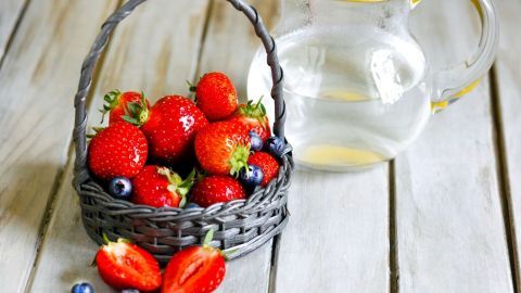 Eine schwarze Korb mit frischen Erdbeeren und Blaubeeren steht neben einem Krug mit heißem Wasser auf einem Holztisch.