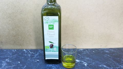 Wunderbar weiche Haut und preiswerte Körperpflege mit nachhaltigem Effekt - baden mit Olivenöl - einfach ausprobieren, es macht Spaß. Eine Flasche Olivenöl steht neben einem Glas mit goldenem Öl auf einem Tisch vor einem neutralen Hintergrund.