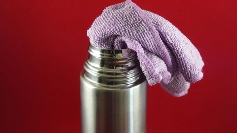 Braunen Belag in der Thermoskanne mit heißem Wasser und Mikrofasertuch entfernen. Eine Thermoskanne wird mit einem lila Mikrofaserhandtuch gereinigt, vor einem roten Hintergrund sichtbar.