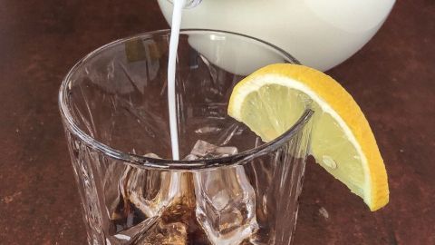 Ein Glas mit dunklem Getränk wird mit heller Milch gefüllt, garniert mit einer Zitronenscheibe, vor einem holzähnlichen Hintergrund.