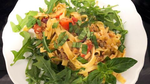 Nudelsalat liegt auf einem weißen Teller, umgeben von frischem Rucola und Kräutern, mit Paprikastücken und gehacktem Basilikum.