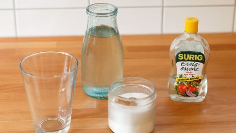 Vase mit heißem Wasser füllen, Zutaten reingeben, über Nacht stehen lassen. Wirkt bestimmt, der Tipp ist von meiner Mutti. Eine Glasvase steht neben einer Wasserkarafe und einem Behälter mit Essig und Salz auf einem Holzuntergrund.