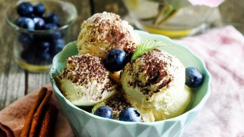 Eine Schüssel mit leckerem Dessert-Eis, dekoriert mit Blaubeeren, steht auf einem Holzuntergrund neben einer Vase mit Pfingstrosen.