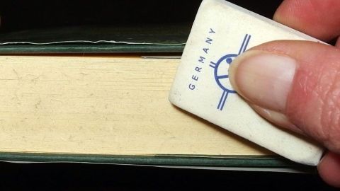 Fingerabdrücke und andere Schmutzspuren auf dem Schnitt eines Buches mit einem Radiergummi, und wenn das nicht hilft, mit feinem Schmirgelpapier entfernen. Ein Buch liegt auf einem Tisch, während eine Hand einen Radiergummi benutzt, um Schmutz vom Buch zu beseitigen.