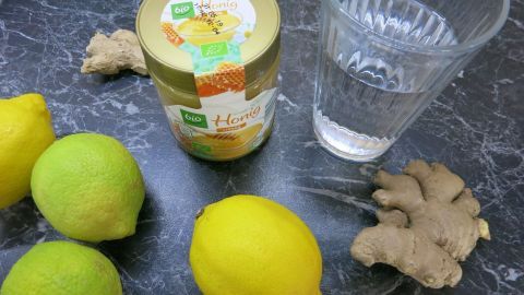 Leckerer und gesunder Ingwerdrink zum Selbermachen. Zusammen mit Honig und Zitrone schmeckt das Getränk einfach himmlisch! Frische Ingwerwurzel, Zitronen und ein Glas Wasser liegen auf einer dunklen Oberfläche zur Zubereitung eines Vitamindrinks.