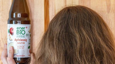 Eine Person hält eine Flasche mit einem Läusemittel über ihrem Kopf, während sie vor einer Holzoberfläche steht.