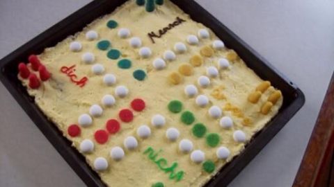 Ein bunter Mensch-ärgere-dich-nicht-Kuchen liegt in einer Backform auf einem Tisch, dekoriert mit essbaren Spielfiguren.