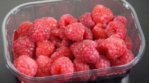 Frische Himbeeren liegen in einer transparenten Plastikschale auf einer dunklen Oberfläche, bereit zum Verzehr.
