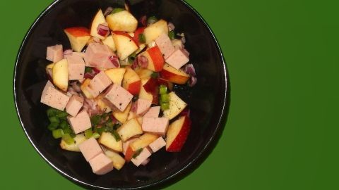 Wurstsalat mit Apfelstücken und Zwiebeln in einer schwarzen Schüssel auf grünem Hintergrund.