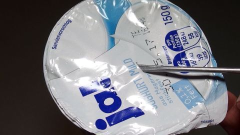 Ein Joghurtbecher wird mit einer Schere geöffnet. Die Umgebung ist dunkel, was den Fokus auf den Joghurt lenkt.