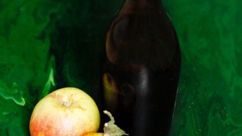 Eine dunkle Flasche Apfelessig steht neben zwei Äpfeln auf einer grünen Fläche, die frisch und lebendig wirkt.
