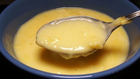 Ein Löffel cremiger Pudding mit einer dicken Haut wird aus einer blauen Schüssel herausgehoben und präsentiert.