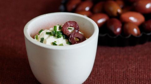 Ein cremiger Feta-Dip in einer weißen Schale, garniert mit Oliven und frischen Kräutern, vor einem Hintergrund mit Oliven.