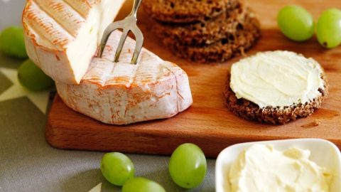 Eine halbe Portion Obatzda liegt auf einem Holzbrett, während eine Gabel den Käse anhebt. Im Hintergrund ergänzen Trauben und Schwarzbrot die Arrangierung.