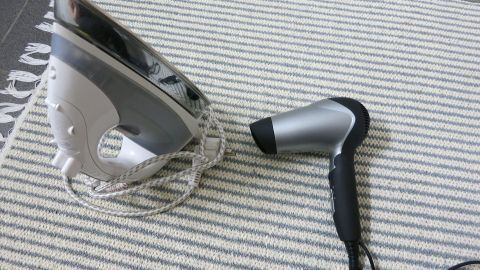 Ein Bügeleisen liegt auf einem Fußbodenbelag, während daneben ein Fön steht. Beide Geräte sind bereit zum Einsatz.