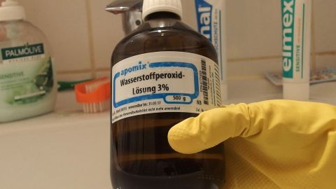 Eine Flasche mit Wasserstoffperoxid wird mit einer gelben Handschuh-Hand gehalten, um Schimmel im Badezimmer zu bekämpfen.