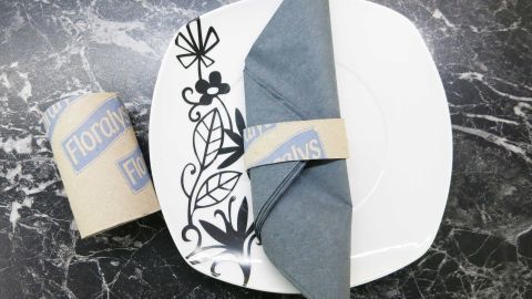 Ein eleganter Serviettenring aus Karton hält eine gefaltete, graue Serviette auf einem stilvollen, dekorierten Teller, platziert auf einer dunklen Oberfläche.