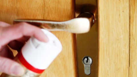 Türschloss klemmt? Einfach etwas Haarspray im eingebauten Zustand hineinsprühen und es lässt sich wieder schließen. Eine Hand sprüht Haarspray auf ein Türschloss, um das klemmende Schloss zu behandeln, in einem holzfarbenen Türrahmen.