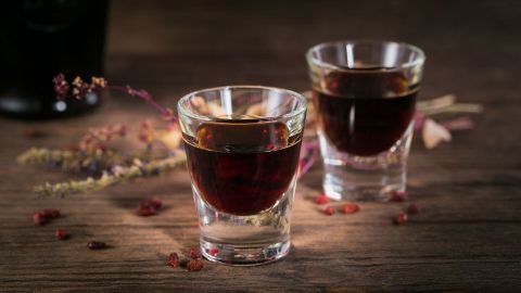 Zwei Gläser mit dunklem Sambuca und Jägermeister stehen auf einer rustikalen Holzoberfläche, umgeben von getrockneten Kräutern.